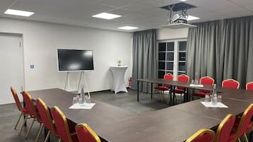 Un salón de conferencias con una larga mesa, sillas rojas, un televisor de pantalla plana, un proyector y una ventana con cortinas.