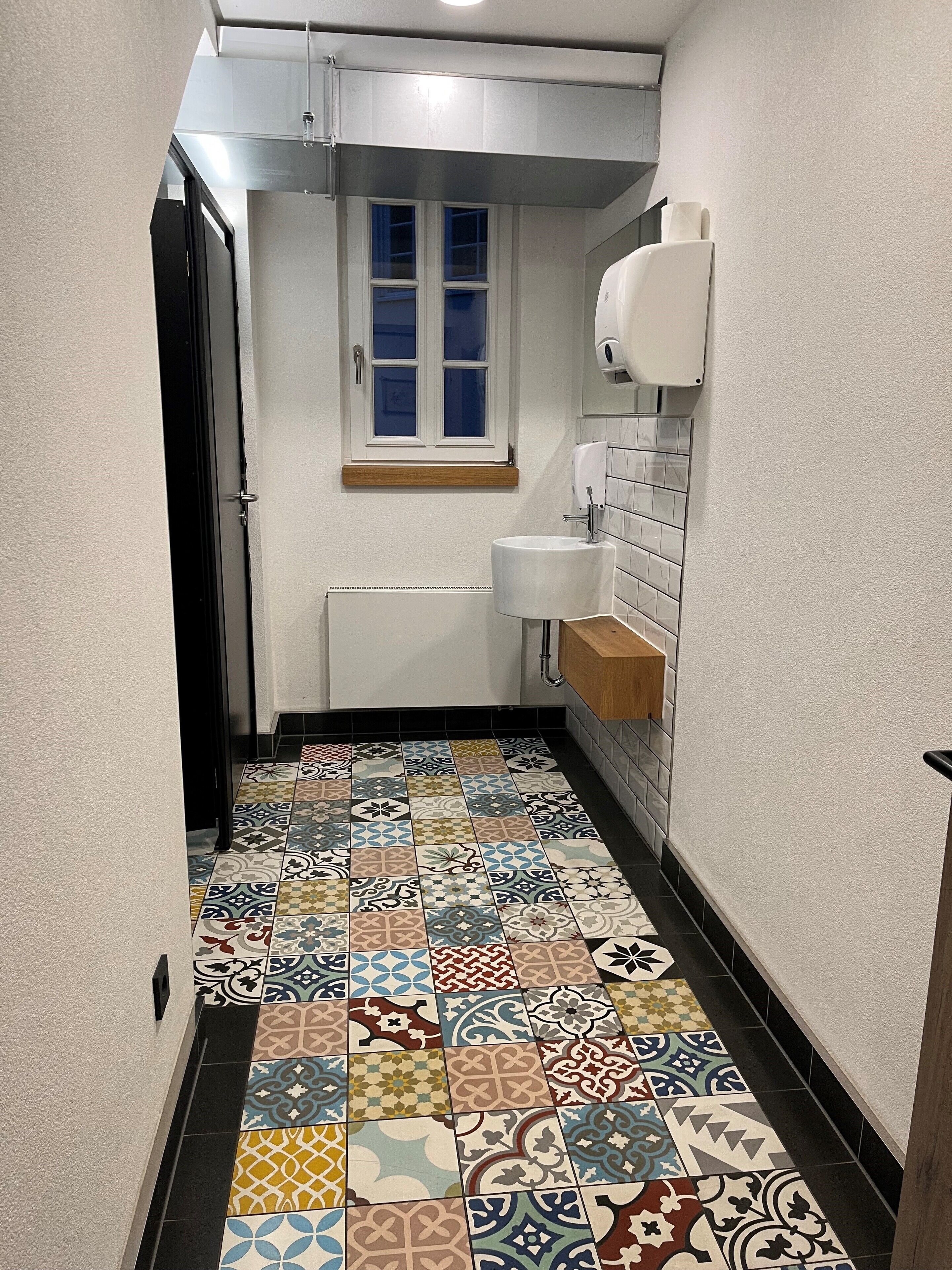A narrow hallway with a tiled floor, a window, a radiator, and a sink.