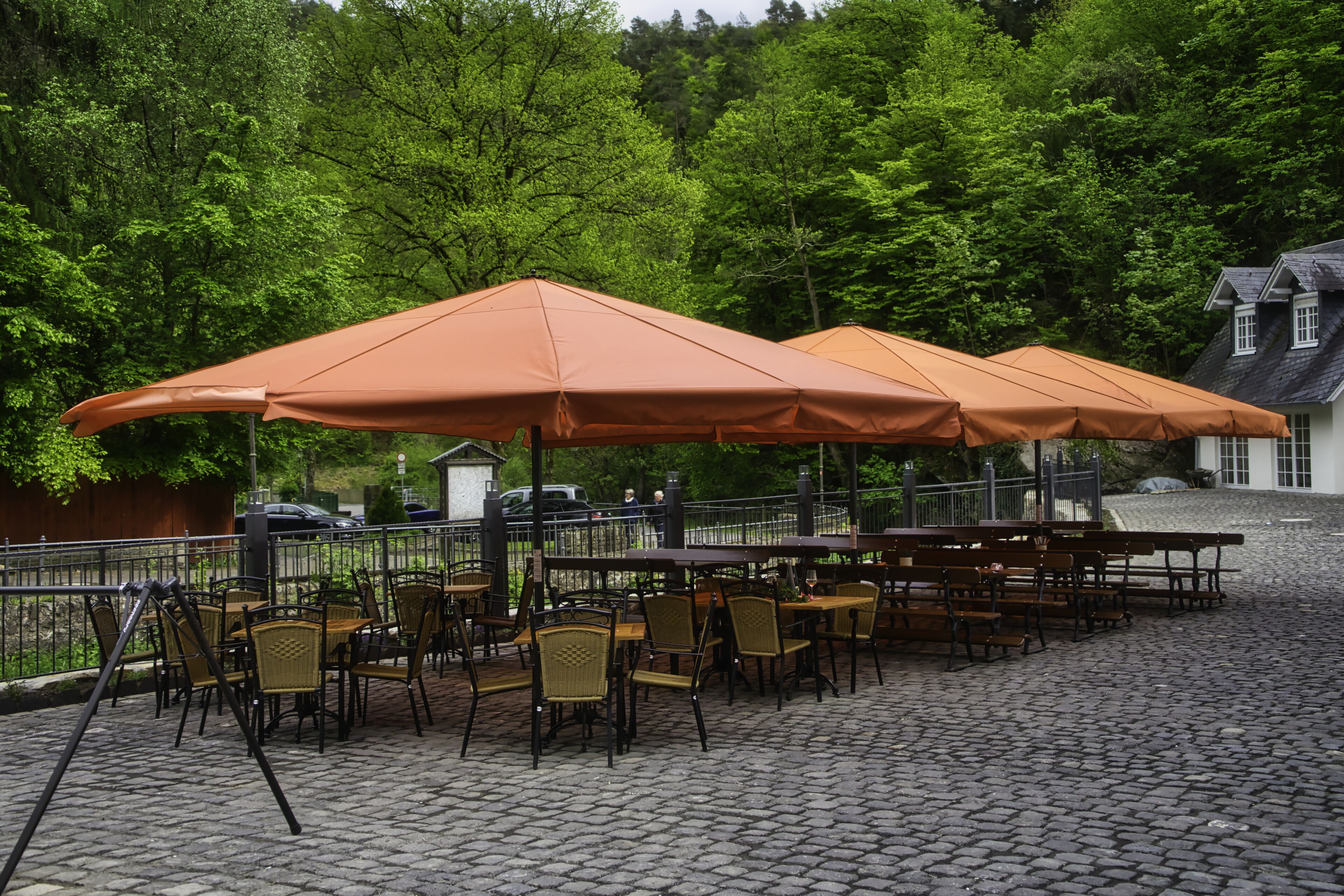 Buitenplaats met zitgedeelte, oranje parasols, kinderaardewerk vloer en een achtergrond van bomen.