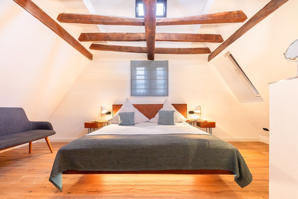 Une chambre avec un plafond en pente, des poutres en bois, un lit recouvert d’une couverture grise, deux tables de chevet avec des lampes et une fenêtre.