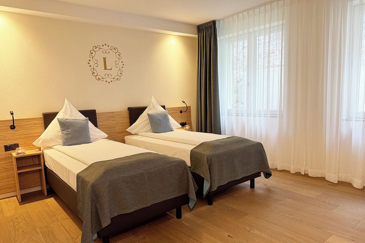 Une chambre d’hôtel avec deux lits, une tête de lit en bois, une table de chevet avec une lampe et une grande fenêtre avec des rideaux légers.