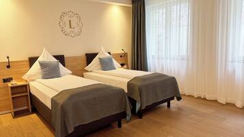 A hotel room with two beds, a wooden headboard, a nightstand with a lamp, and a large window with sheer curtains.