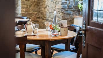 A wooden dining table with chairs, a stone wall in the background, and a door to the right.