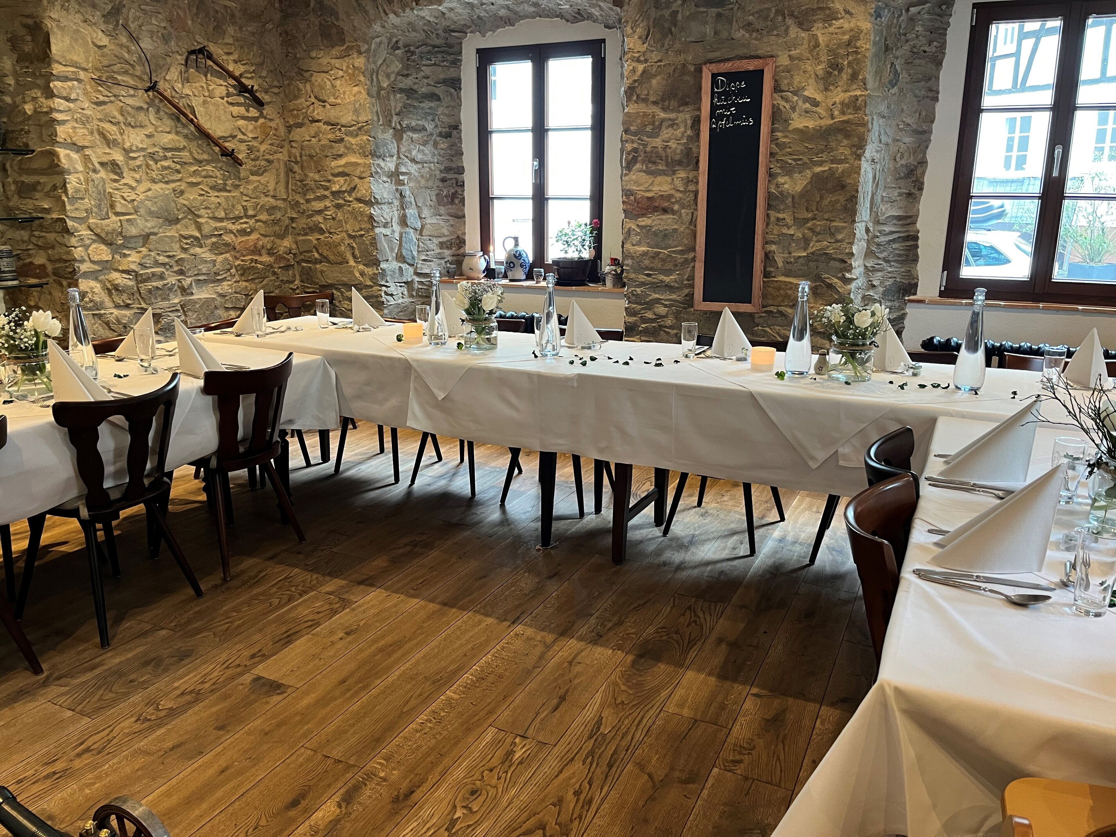 A long dining table set with white tablecloths, wooden chairs, and various table settings including glasses, cutlery, and floral centerpieces.