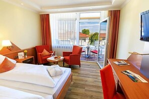 A hotel room with a bed, two red chairs, a small table with a lamp, a TV, and a balcony with a view. - Kurhotel Sonnenhof (Bad Fuessing)