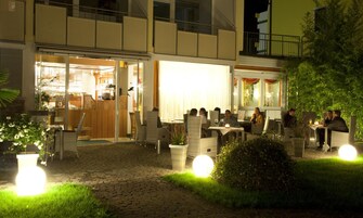 An outdoor dining area with wicker furniture and round tables, surrounded by greenery and a paved walkway.