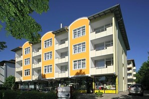 A multi-story building with balconies, a ground-level shop, and a parking area. - Kurhotel Sonnenhof (Bad Fuessing)