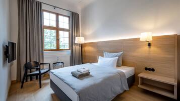 A hotel room with a bed, a desk, a chair, a window with curtains, and two wall-mounted lamps.