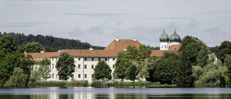 Un edificio grande con una torre y cúpulas destacadas, ubicado junto a un lago tranquilo con árboles densos en primer plano.