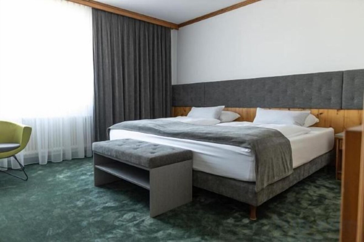A hotel room with a bed, a bench, a chair, and a large window with curtains.