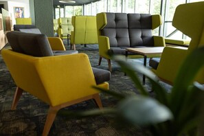 Espaço de convívio moderno com poltronas estofadas em amarelo e cinza, uma mesa central e grandes janelas que proporcionam iluminação natural.