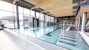 Une piscine intérieure avec des parois en verre transparent, des poutres en bois et un sol carrelé.