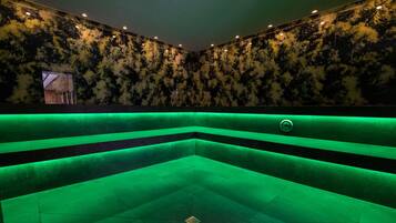Ein Zimmer mit einer Eckbadewanne im grünen Licht, einer Wand mit Muster und Einbauleuchten.