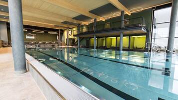 A modern indoor swimming pool with a glass barrier, a mezzanine level, and a staircase leading to a balcony.