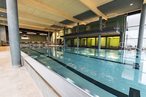 A modern indoor swimming pool with a glass barrier, a mezzanine level, and a staircase leading to a balcony. - Gut Matheshof BW Signature Collection (Rieden)