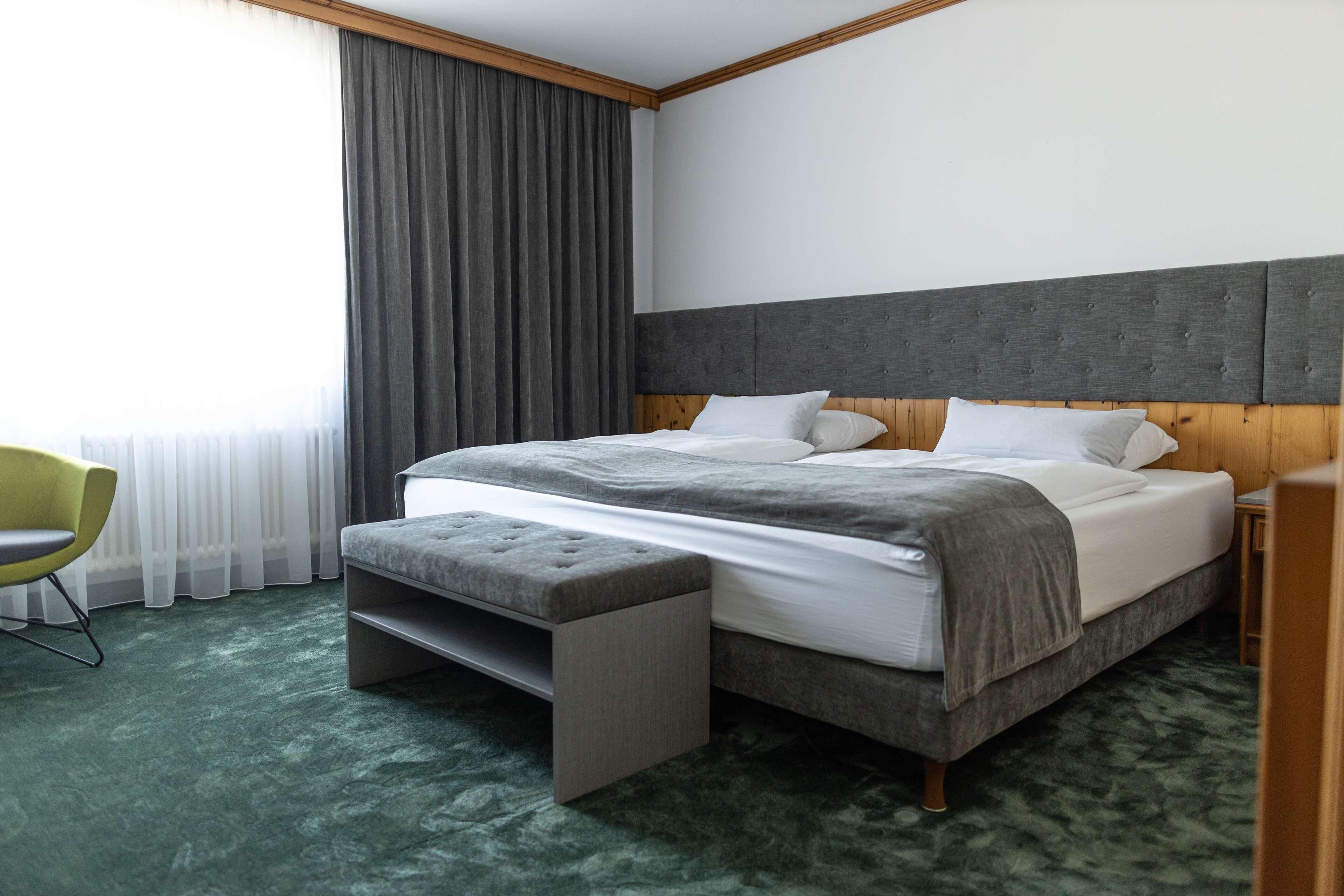A hotel room with a bed, a bench, a chair, and a large window with curtains.