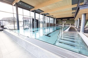An indoor swimming pool with clear glass walls, wooden beams, and a tiled floor. - Gut Matheshof BW Signature Collection (Rieden)