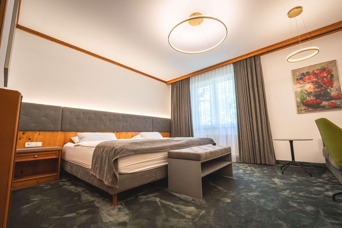 A hotel room with a bed, bedside table, bench, chair, and a painting on the wall.