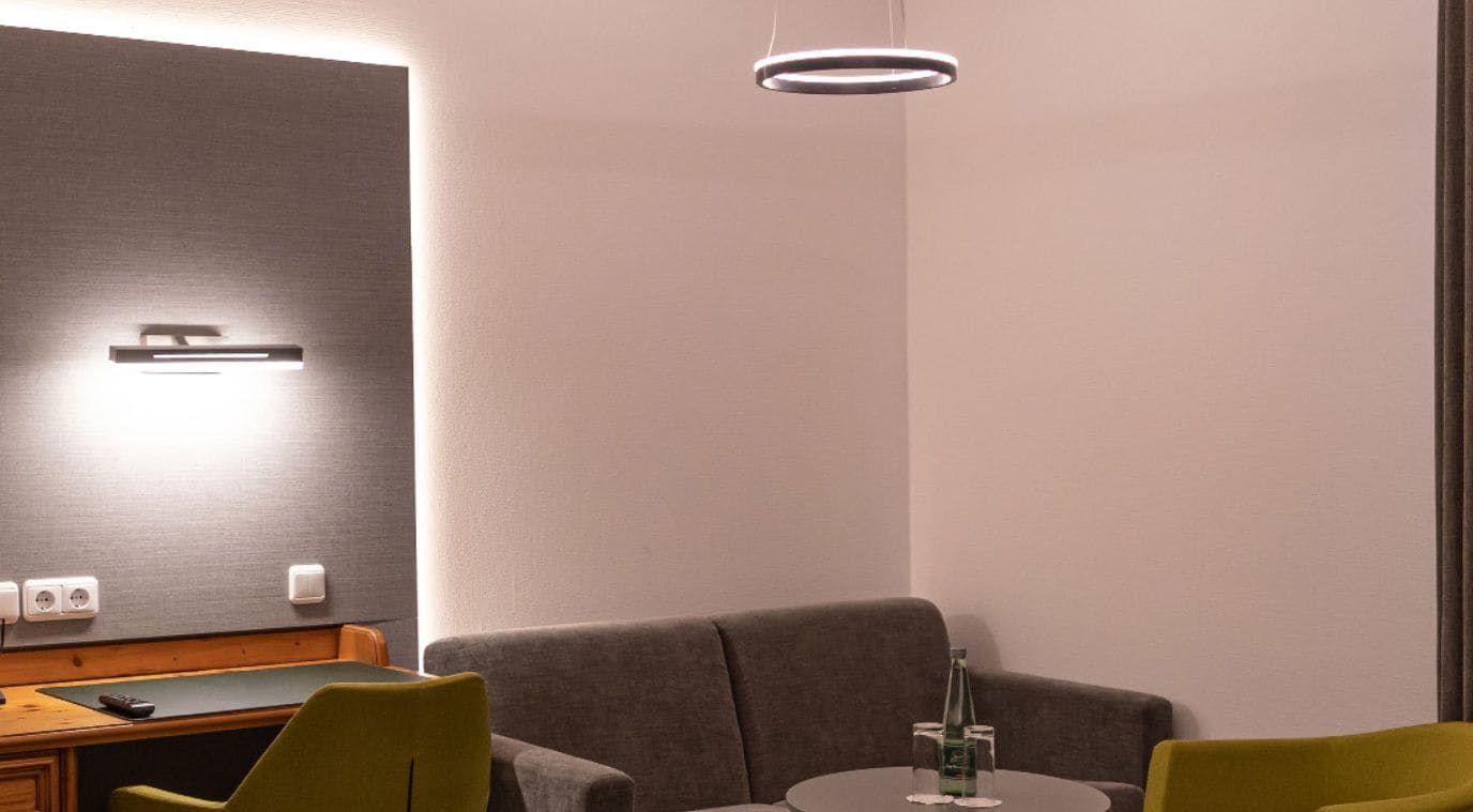Habitación de hotel moderna con un sofá gris, una pequeña mesa redonda con una botella y vasos, un escritorio de madera con un control remoto y una lámpara de pared.