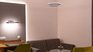 Habitación de hotel moderna con un sofá gris, una pequeña mesa redonda con una botella y vasos, un escritorio de madera con un control remoto y una lámpara de pared.