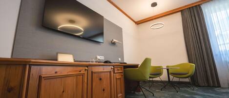 Une chambre d’hôtel avec un bureau en bois, un téléviseur à écran plat fixé au mur, deux fauteuils verts et une grande fenêtre avec des rideaux.