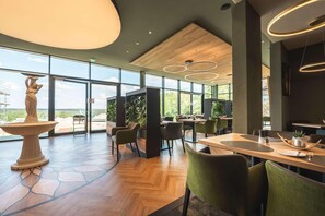 A spacious indoor area with large windows, wooden flooring, green chairs, and a central table with a vase and a plant. - Gut Matheshof BW Signature Collection (Rieden)