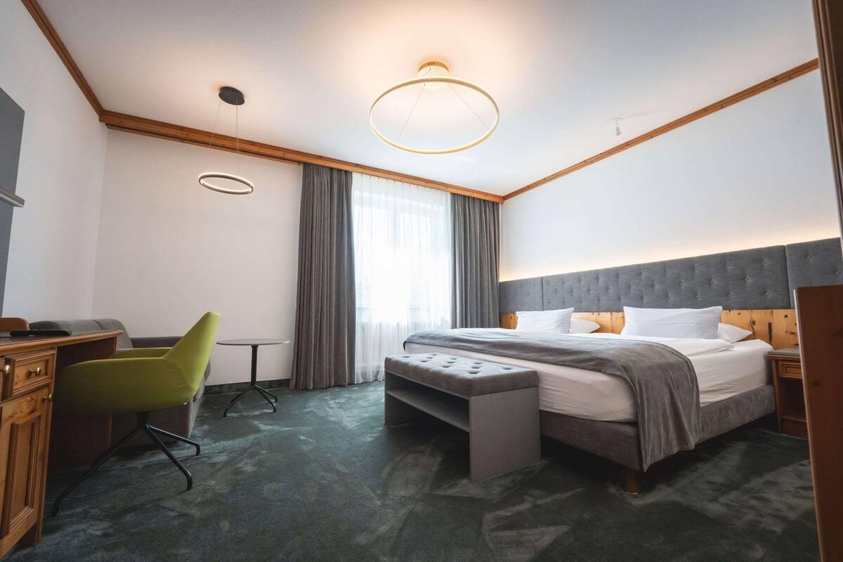 A hotel room with a bed, a desk, a chair, a bench, a bedside table, and a window with curtains.