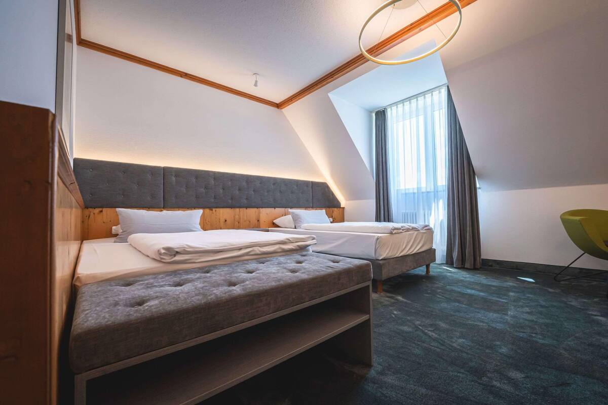 A modern hotel room with two beds, a wooden headboard, a green chair, and a large window.