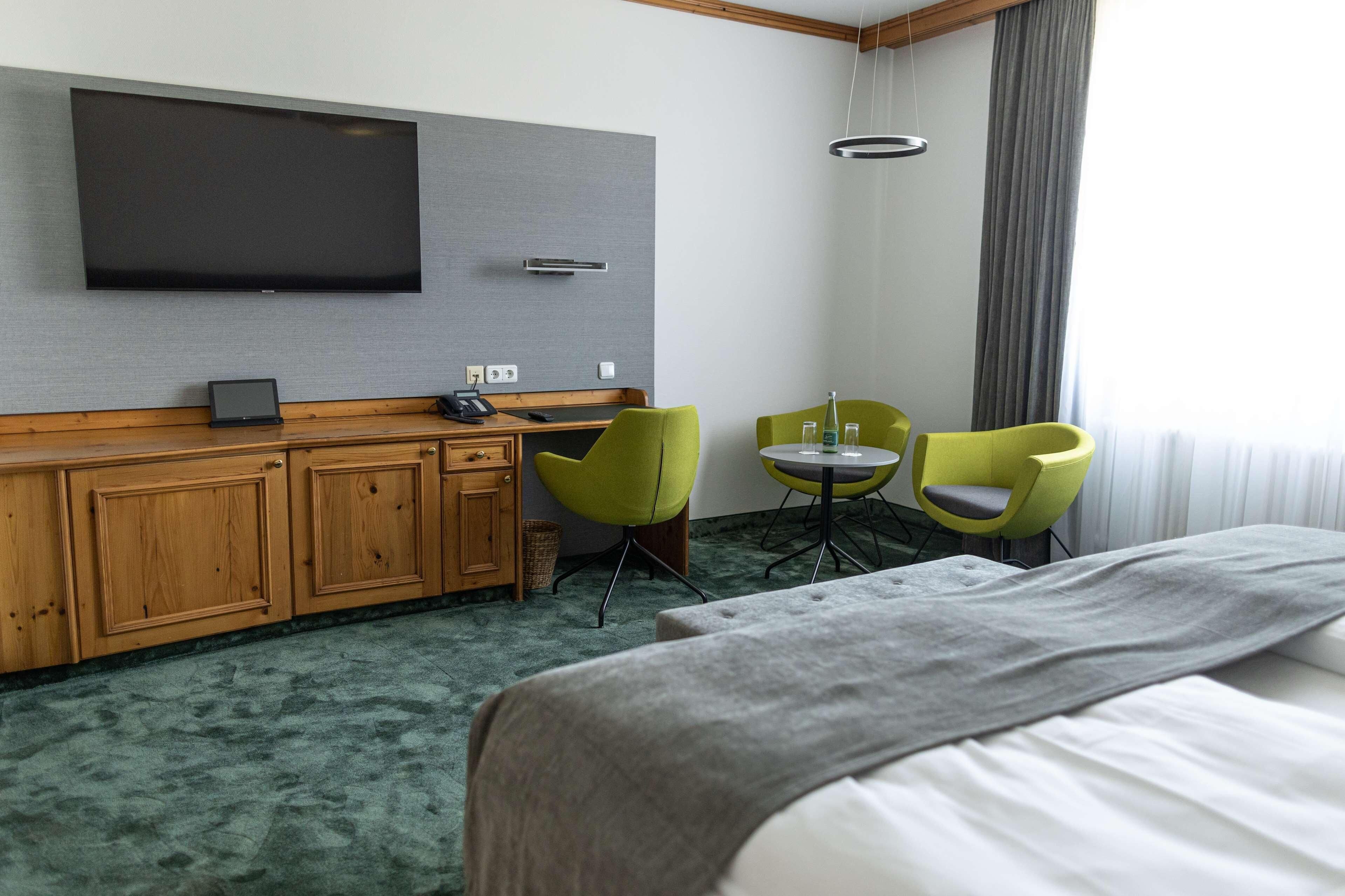 Ein Hotelzimmer mit einem großen Bett, einem an der Wand befestigten Fernseher, einem Holzschrank, zwei grünen Sessel, einem kleinen Tisch und einem Fenster mit Vorhängen.