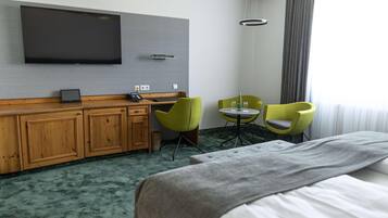 Ein Hotelzimmer mit einem großen Bett, einem an der Wand befestigten Fernseher, einem Holzschrank, zwei grünen Sessel, einem kleinen Tisch und einem Fenster mit Vorhängen.