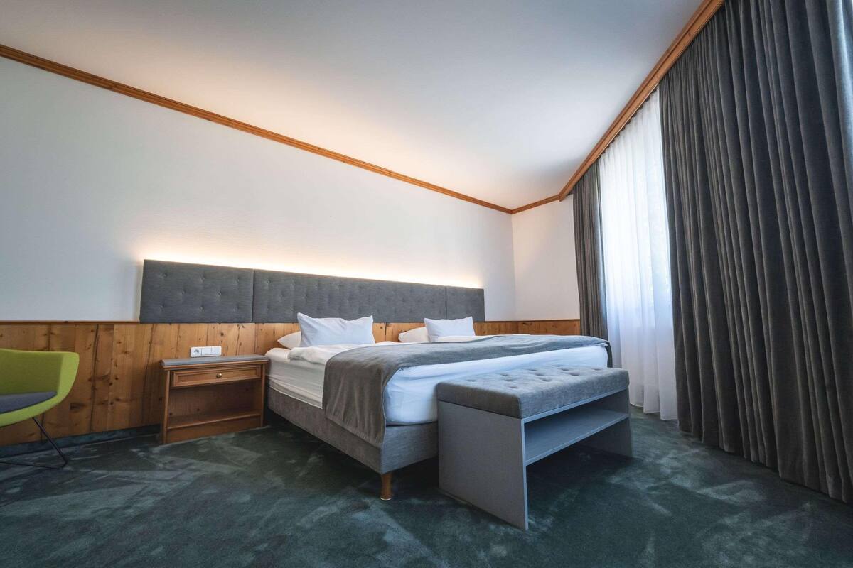 A hotel room with a bed, bedside tables, a chair, and a large window with curtains.