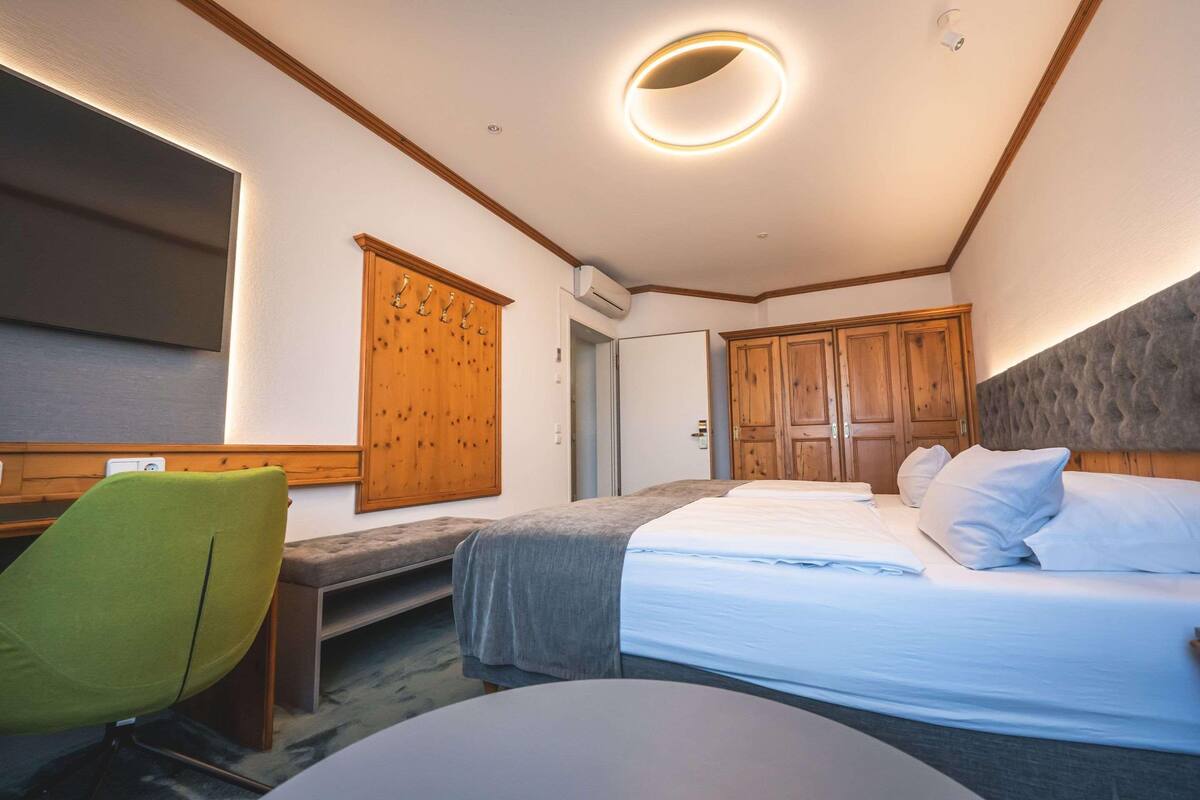 A bedroom with a bed, a bench, a chair, wooden cabinets, and a wall-mounted TV.