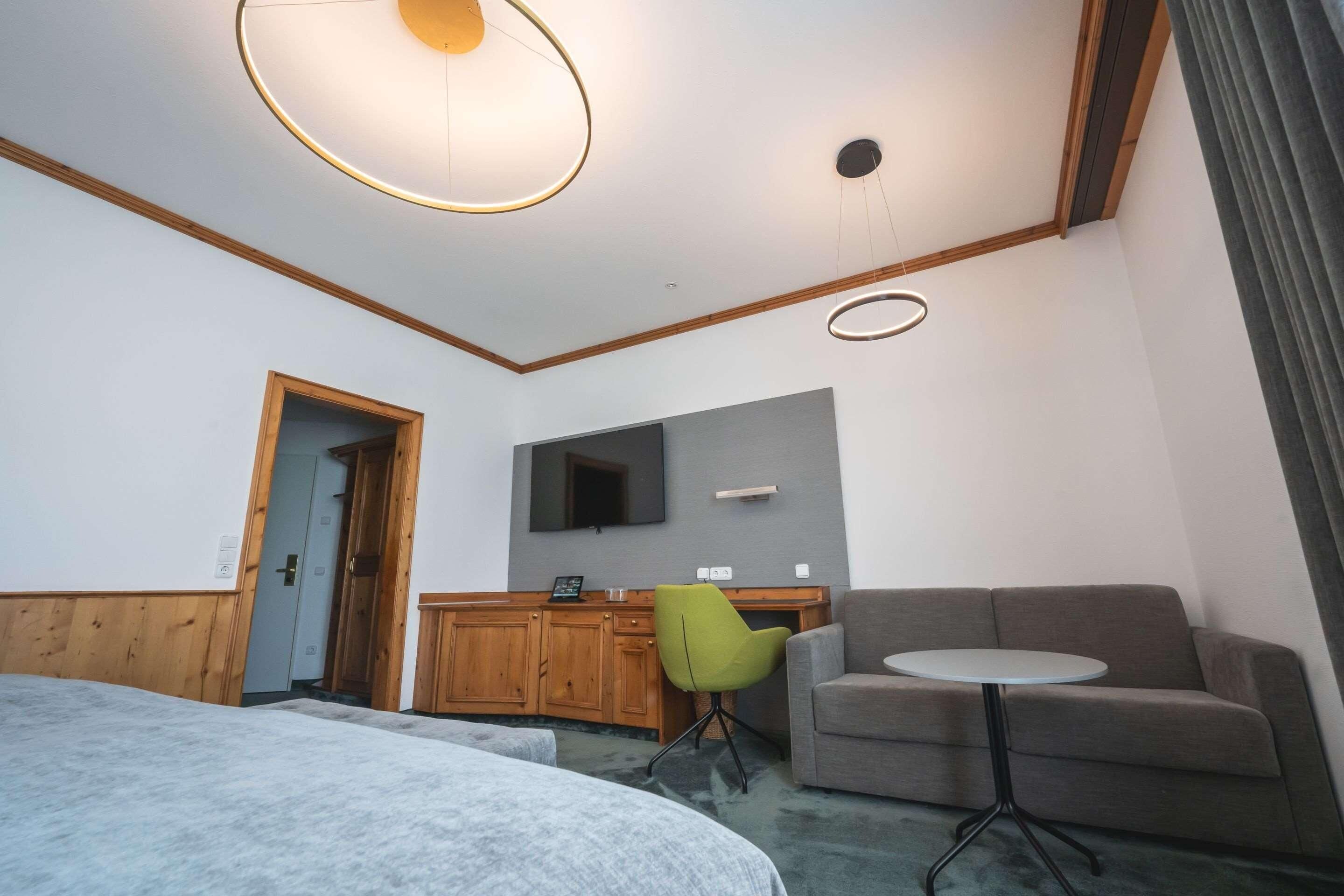 A modern hotel room with a bed, a grey sofa, a small round table, a green chair, wooden cabinets, a flat-screen TV, and a ceiling light fixture.