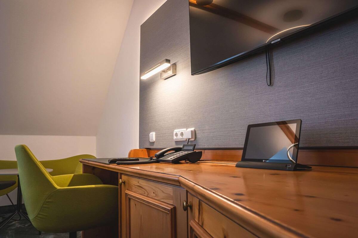 A hotel room with a wooden desk, a green chair, a flat-screen TV mounted on the wall, and a laptop.