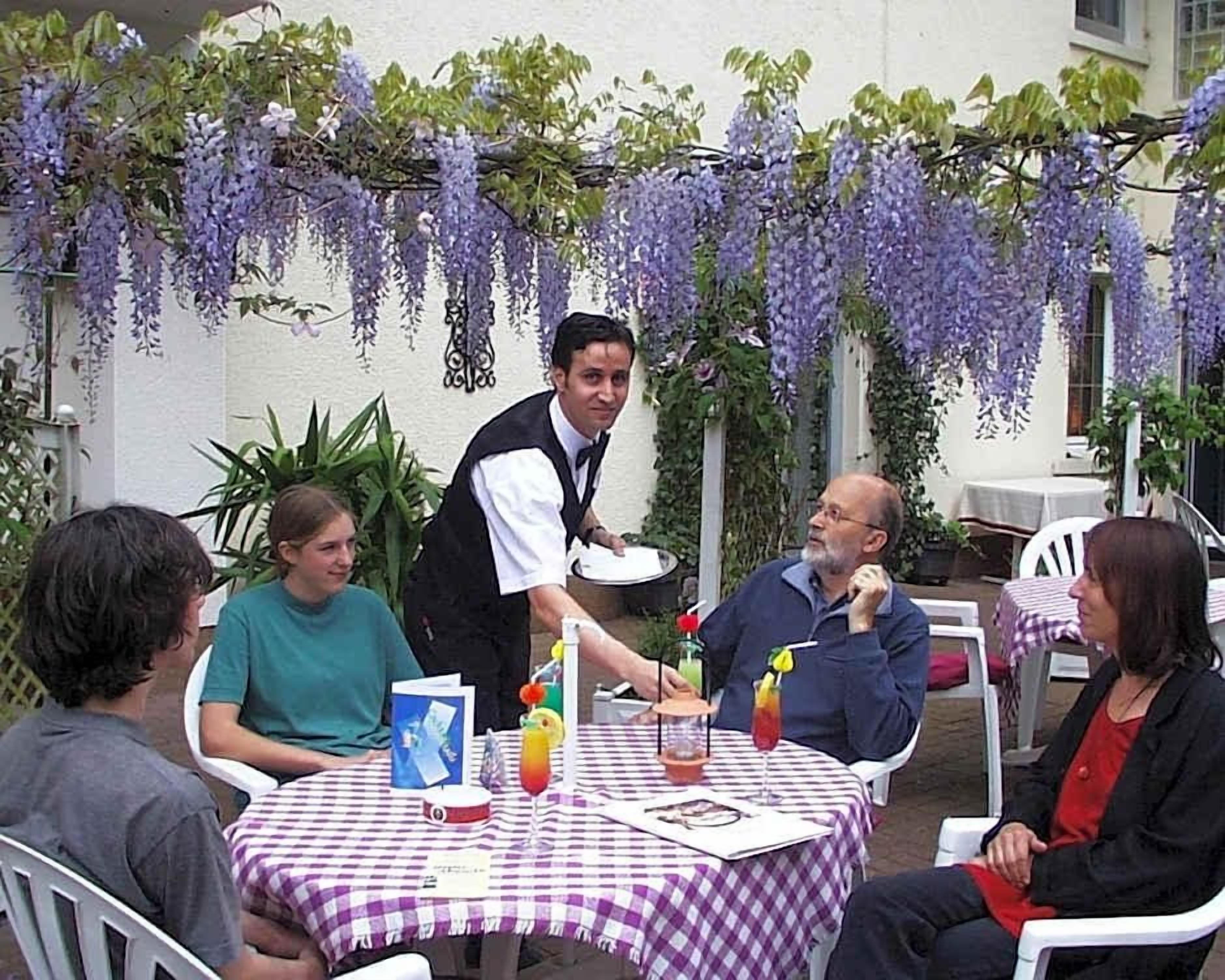 Zona d'exterior amb menjador i taules cobertes amb cambrils a escacs, envoltada de vegetació i enfiladeres de flors morades.