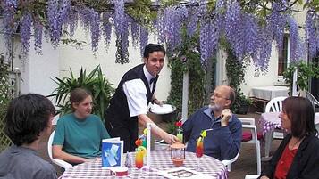 Een terras met eettafels, bedekt met geruit tafelkleed, omgeven door groen en paarse bloeiende wijnranken.