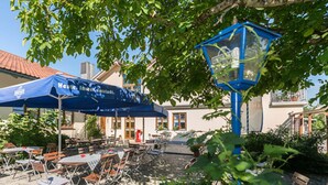 An outdoor seating area with tables and chairs under a blue umbrella, surrounded by greenery and a building with a tiled roof. - Zum Kirchenwirt (Neukirchen vorm Wald)
