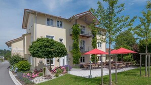 A two-story building with a beige facade, outdoor seating area with red umbrellas, and a well-maintained garden with flowers and shrubs. - Zum Kirchenwirt (Neukirchen vorm Wald)