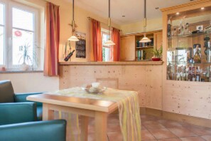 A cozy dining area with a wooden table and chairs, a kitchen with a counter and cabinets, and a window with curtains. - Zum Kirchenwirt (Neukirchen vorm Wald)