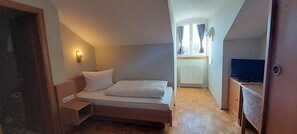 A small, narrow room with a bed, a nightstand, a television, and a window with curtains.