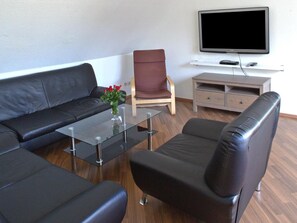 A modern living room with a black leather sofa, a glass coffee table, a wooden chair, and a television on a stand. - Hotel Zum Wersehof (Ahlen)