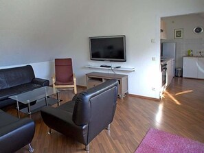 A modern living room with a flat-screen TV, leather sofas, a glass coffee table, and a kitchen area in the background.