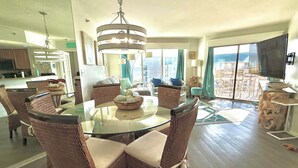 Un vaste espace de vie comprenant un coin repas avec une table en verre entourée de chaises en osier, un lustre et une grande fenêtre offrant une vue sur la ville.