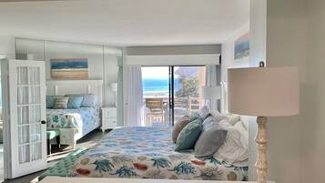 A bedroom with a bed, a nightstand, a lamp, and a view of the ocean through a sliding door.