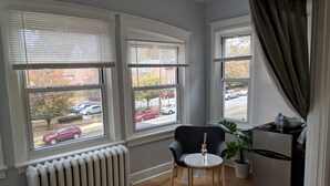 A room with three windows, a radiator, a small table with a bottle, a chair, a refrigerator, and a potted plant. - Lovely, cozy Beacon guest suite with portable kitchen in Squirrel Hill (Pittsburgh)