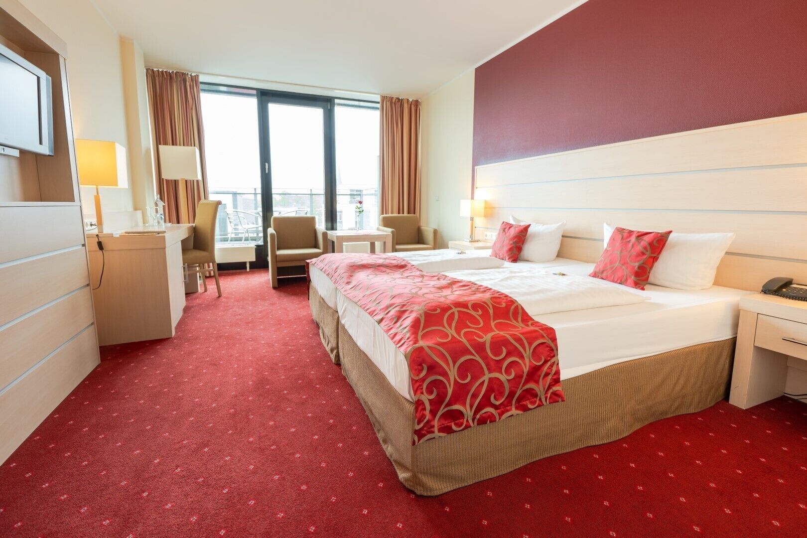 Ein Hotelzimmer mit einem großen Bett, zwei Nachttischen, einem Schreibtisch und einem Stuhl. Ein Fenster mit Vorhängen bietet einen Blick nach außen.