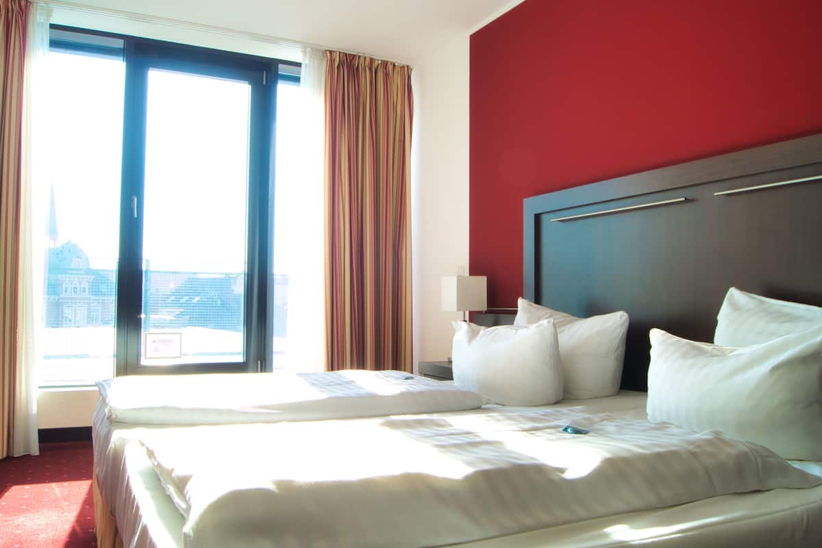 A hotel room with a large bed, a nightstand, and a view of a cityscape through a large window.