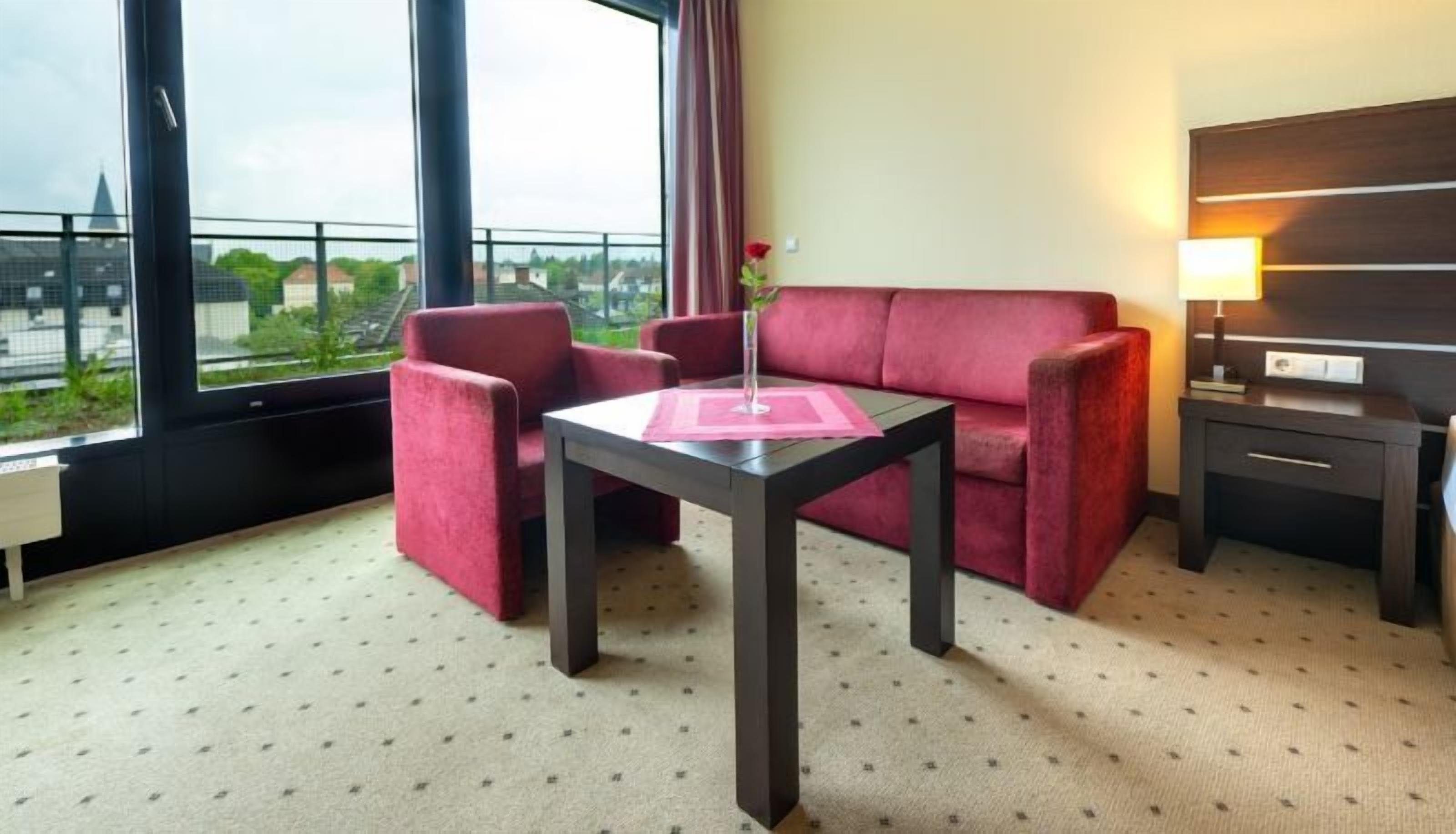 Quarto de hotel com sofá vermelho, uma mesinha com um vaso, uma luminária e vista para prédios através de grandes janelas.