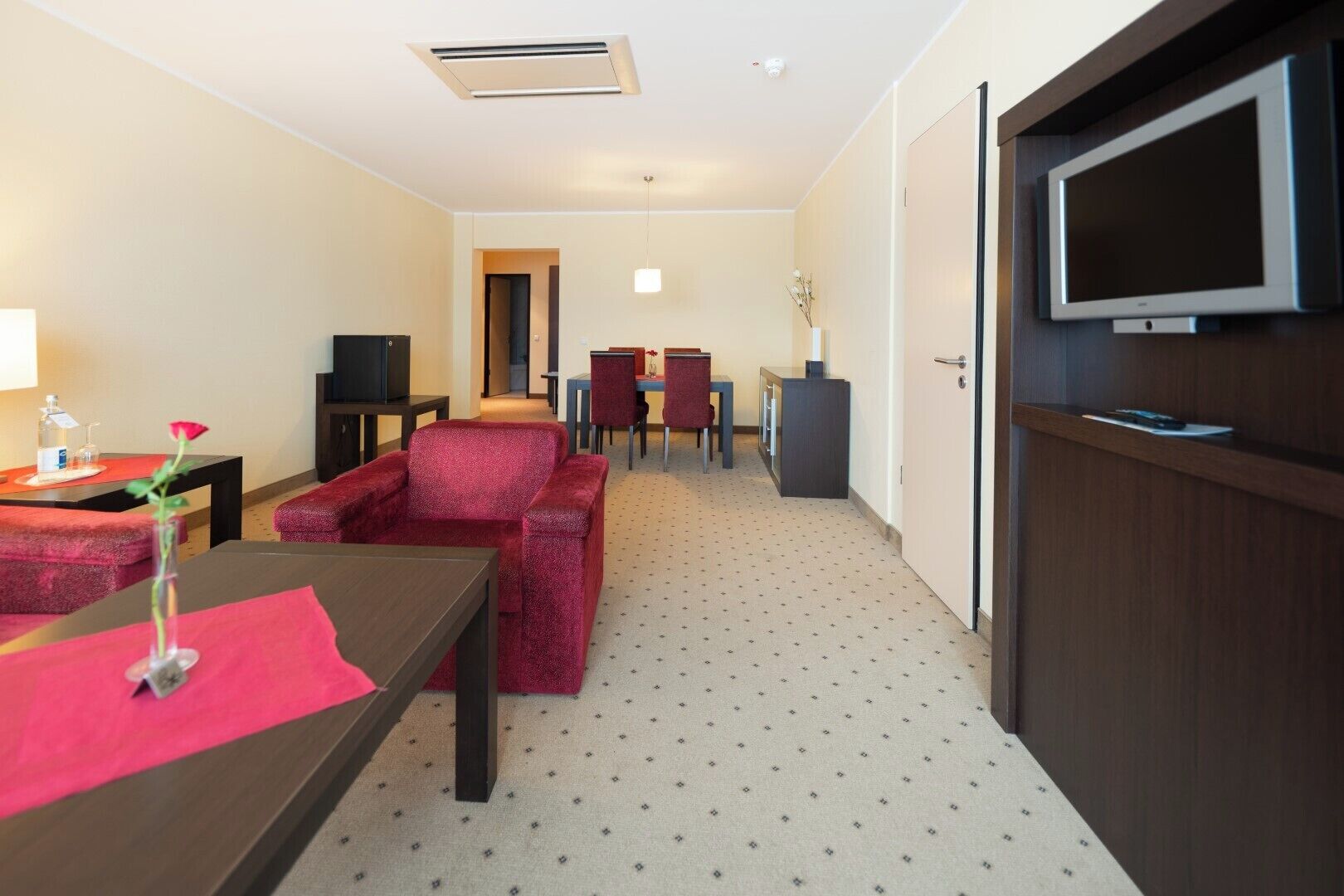 A hotel room with a sofa, a small table with a vase of flowers, a TV mounted on the wall, and a door.