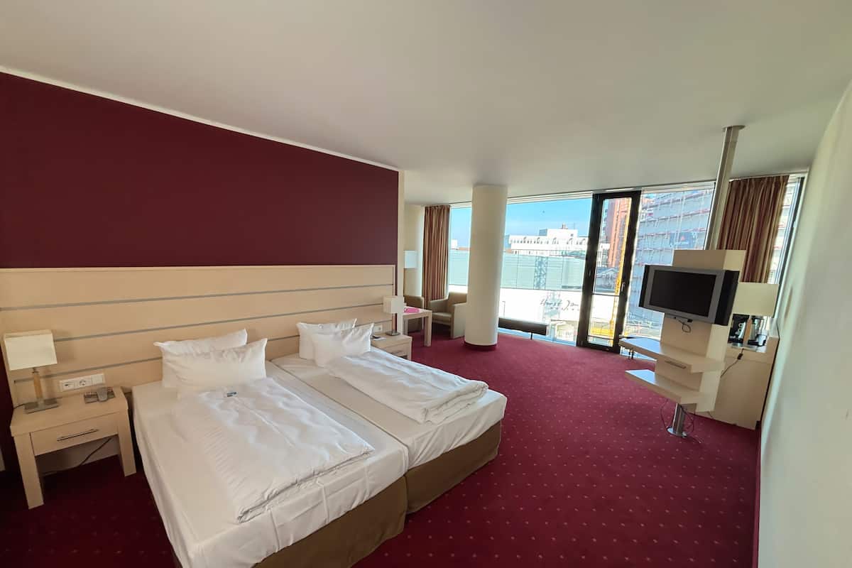 A hotel room with two beds, a desk, a chair, a TV, and a large window offering a city view.